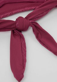 Gros plan d'un nœud noué sur une sangle en tissu bordeaux aux bords scintillants, sur fond gris clair.