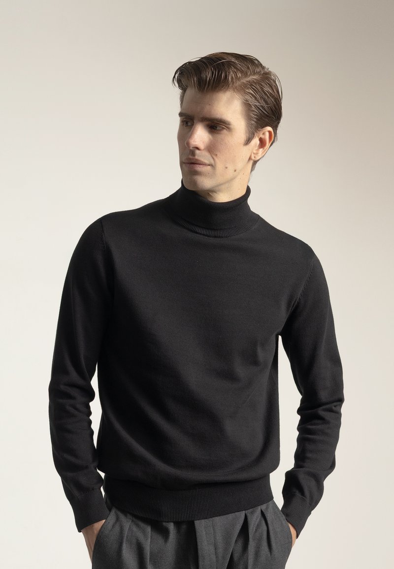 Hombre con el pelo peinado hacia atrás, vistiendo un suéter negro de cuello alto y pantalones grises, de pie con las manos en los bolsillos, mirando hacia un lado.