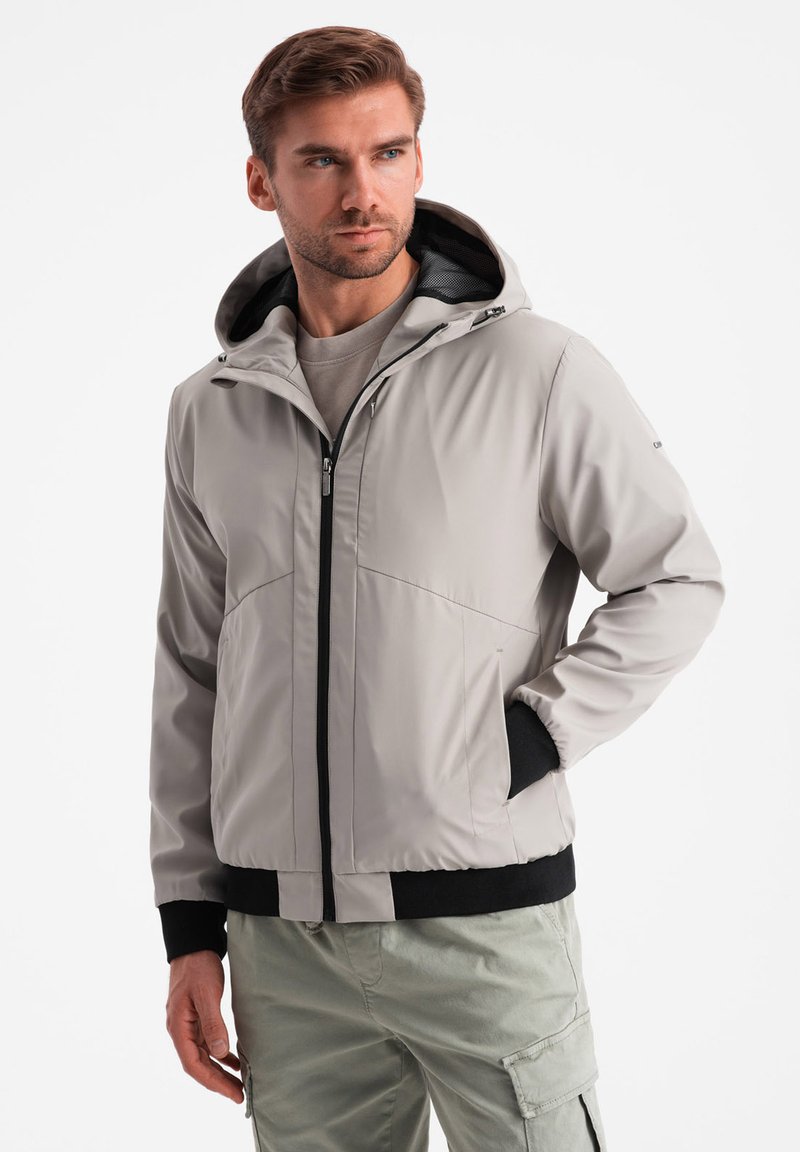Giacca a vento grigio chiaro con cappuccio e zip frontale, dettagli neri ai polsini e all'orlo, in un materiale liscio e impermeabile.