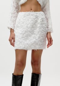 Mini-jupe blanche avec une surface texturée et effilochée. Associée à un haut transparent assorti et portée avec des bottes noires montantes.