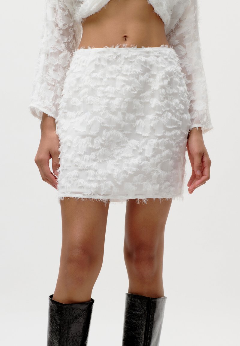 Mini-jupe blanche avec une surface texturée et effilochée. Associée à un haut transparent assorti et portée avec des bottes noires montantes.