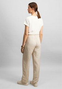 Beige linnedagsbukser med elastisk talje, parret med en hvid t-shirt med korte ærmer. Modellen står med ryggen til foran en grå baggrund.