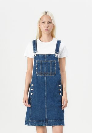 Denim overallkjole med frontlomme, knappdetaljer på siden og rett passform. Brukt over en enkel hvit t-skjorte.