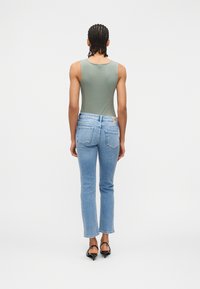 Άτομο με πλεξούδες που φοράει πράσινο τοπ χωρίς μανίκια, ανοιχτό μπλε τζιν και μαύρες σανδάλια με χαμηλό τακούνι, όρθιο με την πλάτη προς το μέρος του θεατή σε λευκό φόντο.