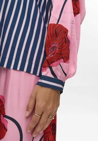 Blouse fluide rose avec imprimé floral rouge, dotée de manches rayées bleu marine et d'un poignet avec un bandeau décoratif. Texture de tissu doux et léger.
