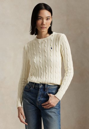Femme portant un pull torsadé crème avec un petit logo et un jean bleu, debout devant un fond neutre uni.