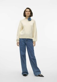 Maglione crema con texture a coste e spalle calate, abbinato a jeans in denim a gamba larga. Un fiore di stoffa blu decora il collo.