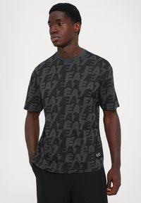 Young man wearing a black and dark gray EA7-branded patterned T-shirt and black pants, standing with one hand in pocket against a plain background.