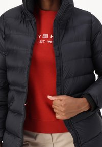 Person wearing a black puffer jacket unzipped, revealing a red sweater with white text, and beige pants, holding jacket near the zipper.
