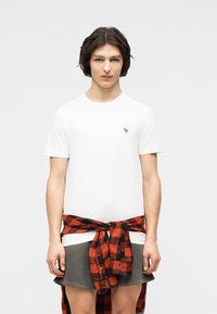 Νεαρό άτομο με κοντά σκούρα μαλλιά, που φοράει λευκό T-shirt και δένει κόκκινο και μαύρο καρό πουκάμισο γύρω από την γκρι φούστα του.