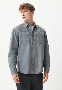 Grå denimskjorte med knapdesign, to brystlommer, lange ærmer og buet søm. Modellen bærer mørke jeans og en hvid T-shirt.