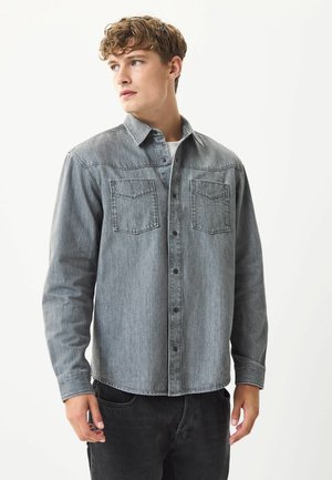 Grijze denim overhemd met een knoopsluiting, twee borstzakken, lange mouwen en een gebogen zoom. Model draagt donkere jeans en een wit T-shirt.