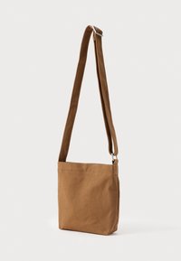 Borsa a tracolla in canvas marrone con tracolla regolabile e fibbia argentata, in posizione eretta su uno sfondo bianco.