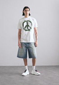 Camiseta de algodón blanca con un símbolo de paz verde. Pantalones cortos de mezclilla azul claro con un desvanecimiento sutil. Zapatillas blancas con acentos en negro y rojo.