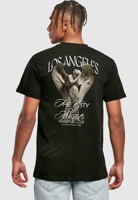 T-shirt en coton noir avec un grand graphique d'un ange enlacé dans des ailes, avec le texte "LOS ANGELES" au-dessus et un slogan en dessous en blanc.