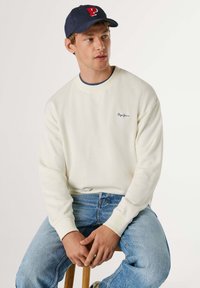 Jonge man met een marineblauwe pet met een rode "P", een wit Pepe Jeans sweatshirt en lichtblauwe spijkerbroek, zittend op een houten kruk tegen een witte achtergrond.