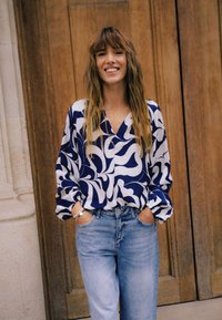 Sinine ja valge mustriga pluus V-kaeluse ja pikkade varrukatega, kombineeritud helesiniste teksadega. Taustal puituksed.