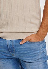 Μπεζ ριγωτό πλεκτό πουλόβερ με ριγέ στρίφωμα, σε συνδυασμό με μπλε τζιν παντελόνι με ξεθωριασμένη υφή. Χέρι που ακουμπάει στην τσέπη.