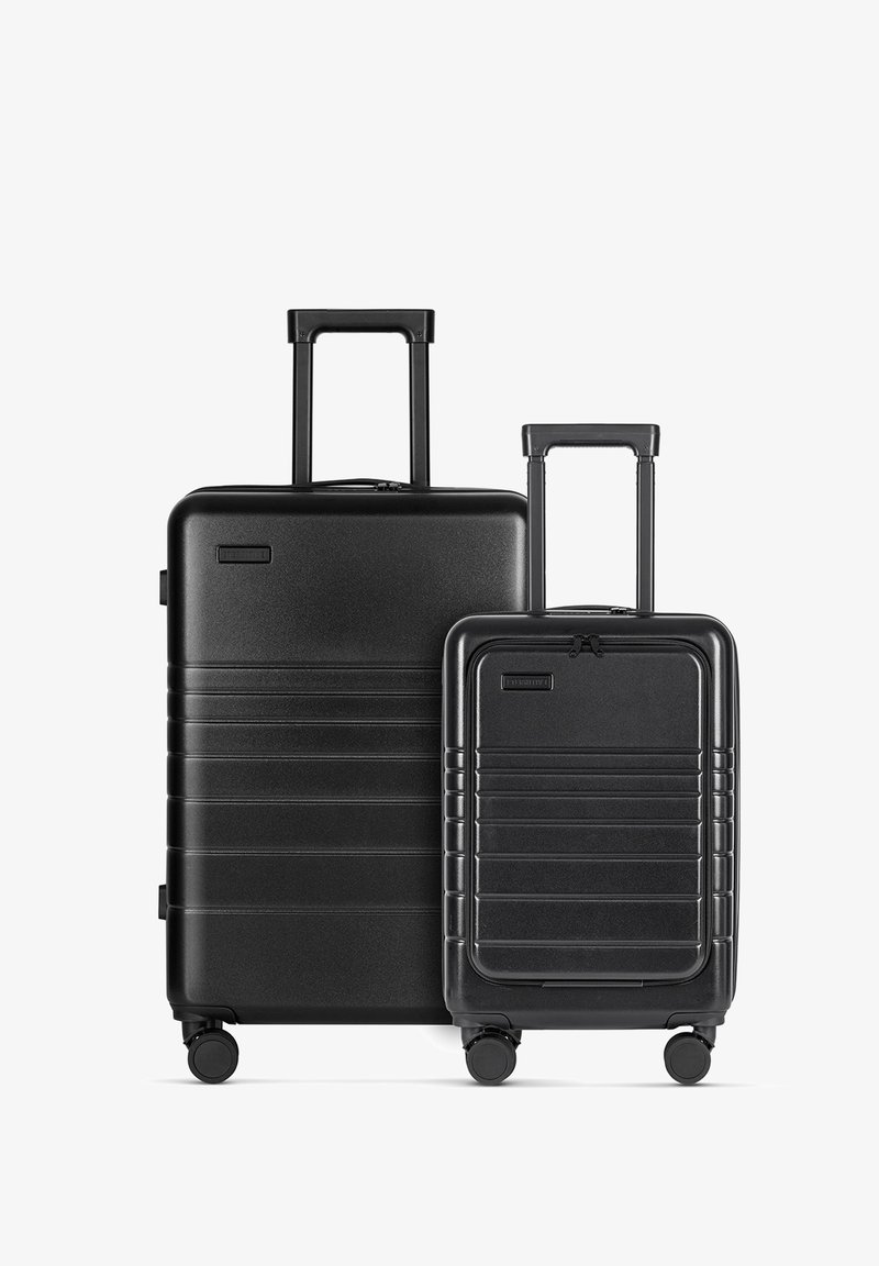 Twee zwarte harde kofferbakken, de ene groter met een telescopische handvat en wielen, de andere kleiner, beiden met ribbelontwerpen en ritsen.