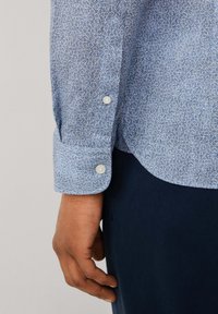 Camisa de patrón azul con diseño floral, de manga larga, con puños abotonados y textura suave. El cuello es ligeramente curvado.