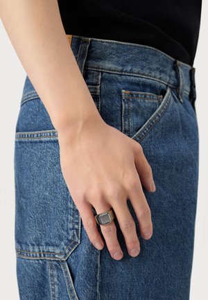 Metallring med en firkantet sort sten, båret på en hånd nær en denimlomme. Jeansene er en falmet blå med synlig syning og tekstur.