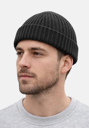 Mann mit kurzem Bart, der eine gerippte schwarze Strickmütze und ein hellgraues Rundhalsshirt trägt, blickt nach links vor einem schlichten hellen Hintergrund.