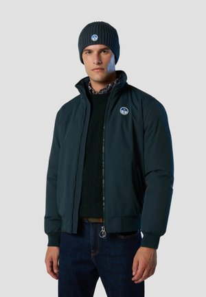 Mann trägt eine marineblaue Mütze und Jacke mit passenden runden Logoaufnähern, schwarzen Pullover, karierten Kragen und dunkle Jeans vor schlichtem Hintergrund.