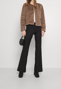 Bruine jas van imitatiebont met een doorgestikt ontwerp, zwarte broek met wijde pijpen en een kleine zwarte handtas. Soepele textuur, knoopsluiting en lange mouwen.