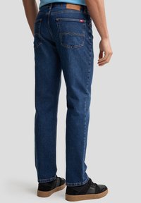 Dunkelblaue Jeans mit geradem Bein, ausgestattet mit zwei Gesäßtaschen und hellbraunem Nähen. Getragen mit schwarzen Sneakers und einem hellblauen Hemd.
