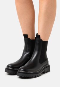 Bottes chelsea en cuir noir avec panneaux élastiques sur les côtés, semelle en caoutchouc texturé et coutures blanches contrastantes le long du bord.