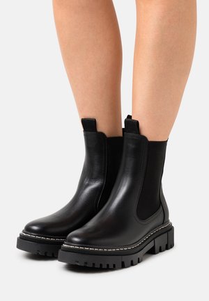 Black leather chelsea boots with elastic side panels, textured rubber sole, and contrasting white stitching along the edge.