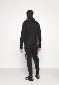 Man med dreadlocks iklädd en svart träningsoverall med vita ränder, stående vänd mot en plain vit vägg.