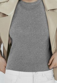 Person wearing a beige jacket over a fitted gray ribbed short-sleeve top tucked into white pants, hands adjusting the jacket sides.