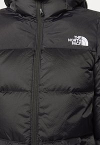 Doudoune matelassée noire avec fermeture éclair à l'avant et logo blanc « The North Face » sur la poitrine gauche.