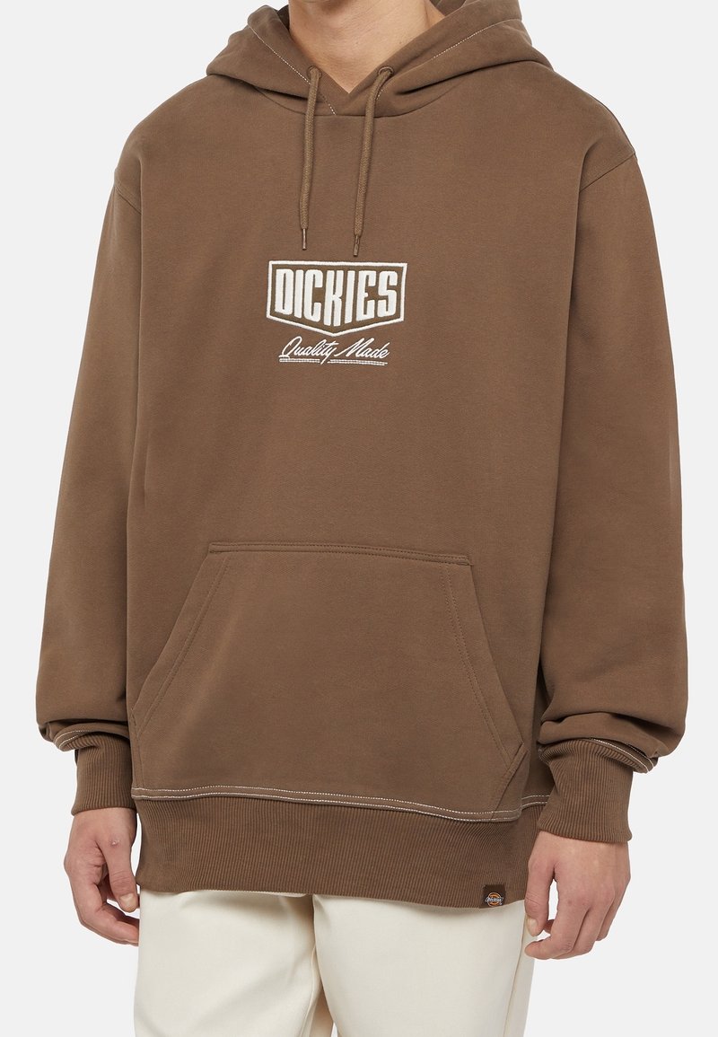 Personne portant un sweat à capuche Dickies marron avec une poche avant et un texte blanc "Dickies Quality Made,", associé à un pantalon de couleur claire.