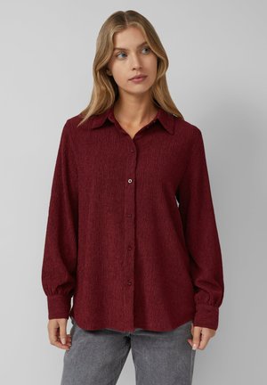 Vrouw met lang blond haar, gekleed in een textuur rimpel bordeauxrode overhemd met knopen en een grijze spijkerbroek, staande tegen een effen lichtgrijze achtergrond.