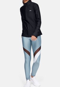 Svart zip-jacka med hög krage; kombinerad med ljusblå leggings som har svart nät och vita accenter i ett chevronmönster. Vita sneakers.