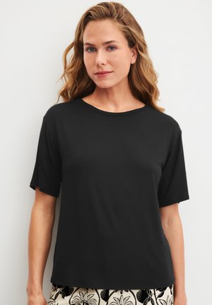 Vrouw met golvend blond haar, gekleed in een eenvoudige zwarte korte mouwt-shirt en zwart-witte, patroonbroek, staand tegen een witte achtergrond.