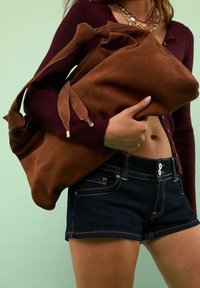 Brun semsketotebag med blød tekstur, justerbare stropper og diskrete guldaccents, holdt op imod en mintgrøn baggrund.