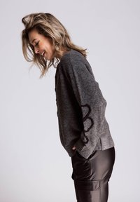 Grauer Wollpullover mit lockerem Schnitt, schwarzen Akzenten an den Ärmeln und einem Etikett an der Seite; kombiniert mit schwarzen Lederhosen.