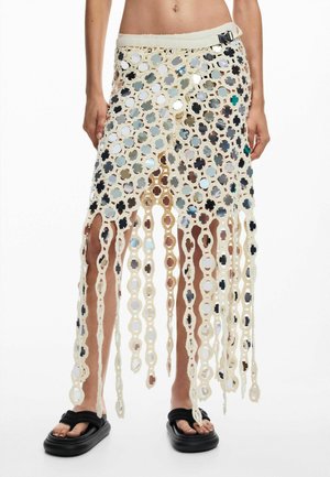 Žena nosi béžovú háčkovanú sukňu s visiaciami slučkami pripomínajúcimi reťaze a pripevnenými malými okrúhlymi zrkadielkami, v kombinácii s čiernymi sandálmi.