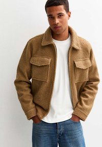 Bruine, gestructureerde fleece jas met een kraag, twee voorzakken en een ritsluiting. Gedragen over een witte blouse, gecombineerd met blauwe jeans.