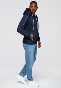 Mężczyzna w granatowej bluzie z kapturem, jasnoniebieskich dżinsach i białych sneakersach, stojący z jedną ręką w kieszeni, patrzący na bok na tle jednolitego tła.