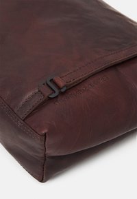 Pochette en cuir brun avec une texture lisse, dotée d'une fermeture éclair et d'un accent noir. Détails cousus le long du bord.