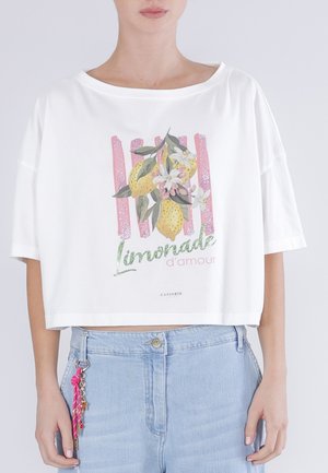 Kvinde iført hvid cropped T-shirt med citron- og blomsterdesign samt teksten "Limonade d'amour", kombineret med lyseblå jeans og nøgleringsvedhæng.