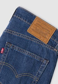 Μπλε τζιν παντελόνι με καφέ δερμάτινο ταμπελάκι που φέρει το λογότυπο της μάρκας, ραμμένες ραφές και κόκκινη ετικέτα στην πλαϊνή τσέπη.