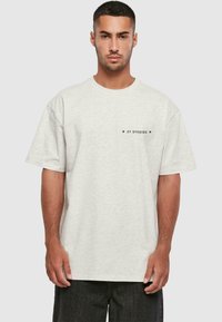 Šviesiai pilkas per didelis t-shirt'as, pagamintas iš minkštos medvilnės, su apvalia iškirpte ir mažuoju juodu logotipu kairėje pusėje.