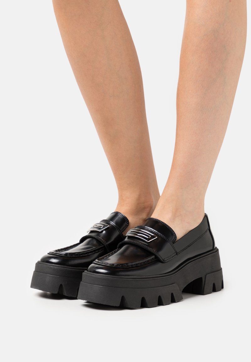 Mocassins noirs avec semelles en caoutchouc épaisses et texturées, finition en cuir brillant, et une bande argentée en travers du cou-de-pied. Design massif.