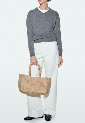 Persona che indossa un maglione grigio, pantaloni bianchi e scarpe nere, che tiene una borsa tote di paglia intrecciata di media grandezza per i manici.