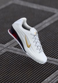 Bijela sportska cipela s teksturiranim gornjim dijelom, zlatnim Nike logom, sivim vezicama i crvenim detaljima na potplatu. Sadrži jastučić s zračnom jedinicom za dodatnu udobnost.